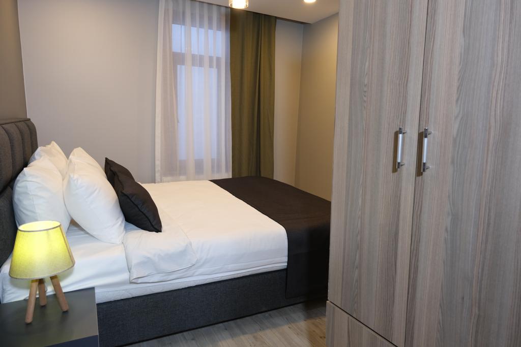 A cozy sleeping area is featured with a spacious bed dressed in white linens and dark accents. A bedside lamp casts a gentle light, and a wardrobe stands nearby. Natural light filters through a window dressed with sheer curtains, creating a calm atmosphere.