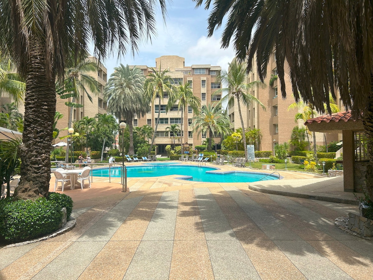 A serene pool area is surrounded by lush palm trees and well-maintained gardens. Lounge chairs are positioned along the water's edge. The spacious pool is complemented by a sun terrace, providing a calming environment for relaxation.