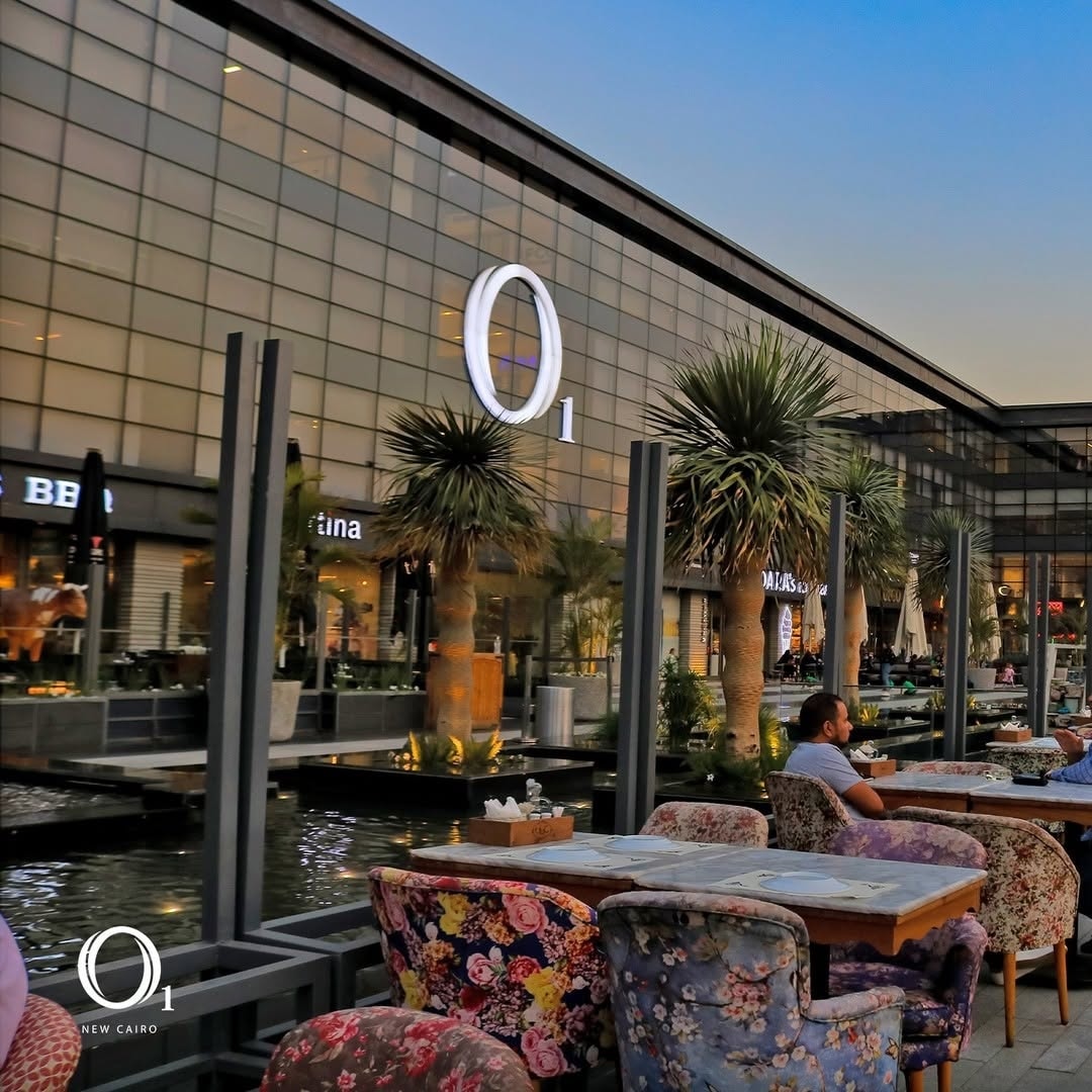 An outdoor dining area at O1 Mall is displayed, featuring colorful floral chairs and wooden tables. Tall palm trees provide greenery, with softly lit seating creating a relaxed atmosphere. Guests can be seen enjoying their meals under the evening sky, enhancing the inviting environment.