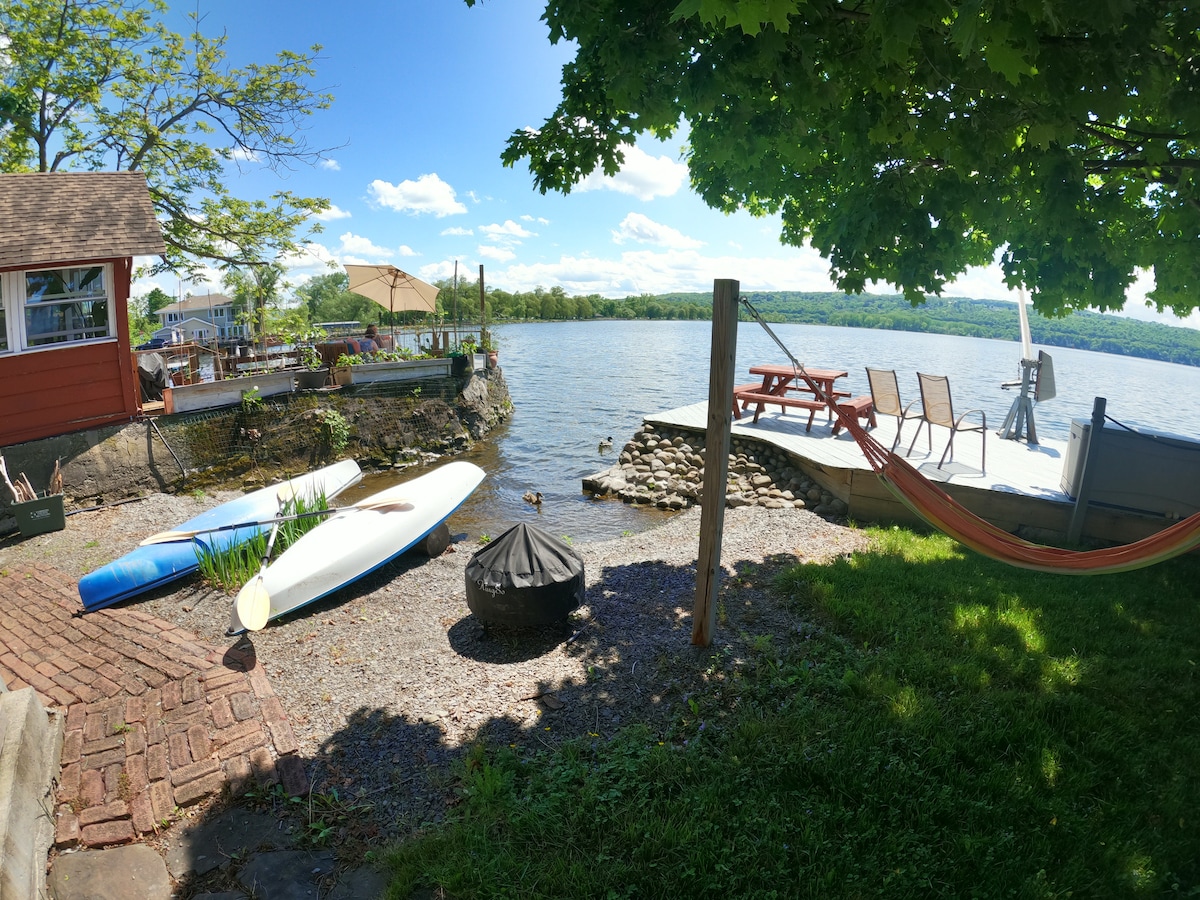 The lakefront area features a grassy space and a stone pathway leading to the water's edge. Two kayaks and a paddleboard rest on the shore. A small firepit stands on gravel, with a picnic table and seating area visible on the deck overlooking the lake.