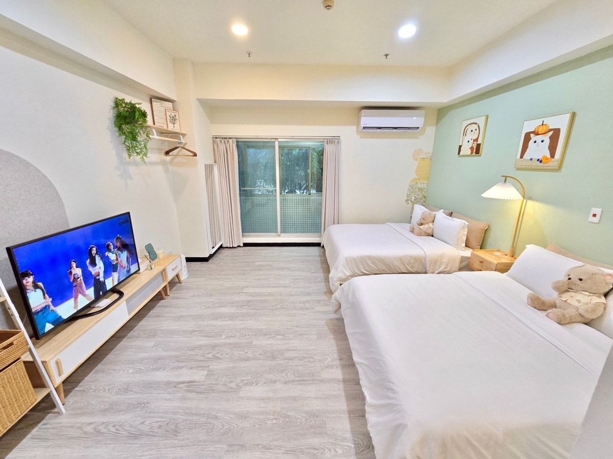 The room features two spacious beds dressed in white linens, complemented by a framed artwork on the green accent wall. A large television stands on a light wood console. Natural light filters through sheer curtains, illuminating the space with a soft glow.