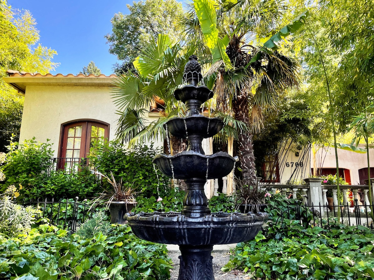 A tiered black fountain is surrounded by lush greenery, including tropical plants and palm trees. The villa's stucco exterior with a tile roof can be seen in the background, adding to the tranquil atmosphere of the garden.