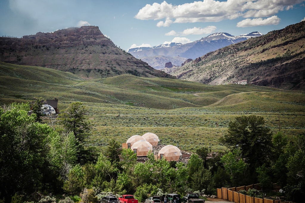 Three geodesic domes are nestled among lush greenery, with rolling hills and mountains in the background. The landscape is dotted with trees and offers scenic views of the distant snow-capped peaks and blue sky, creating a harmonious connection with nature.