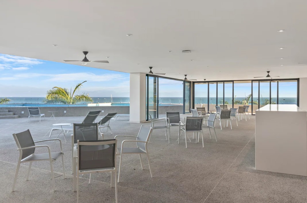 An open space is presented with multiple seating arrangements, featuring white metal chairs facing large glass windows. The ocean view is visible through the expansive windows, with natural light illuminating the area. Ceiling fans hang above, enhancing airflow in the room.