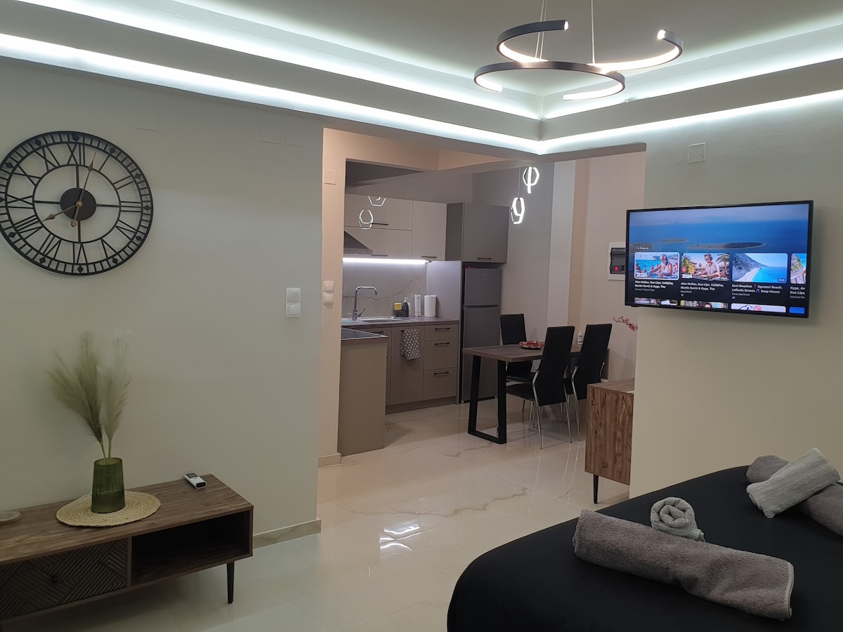 A modern living area features a cozy sleeping space with a neatly arranged bed. A wall clock is positioned above a wooden console table, and a sleek dining area is visible in the background. Soft lighting enhances the overall ambiance.