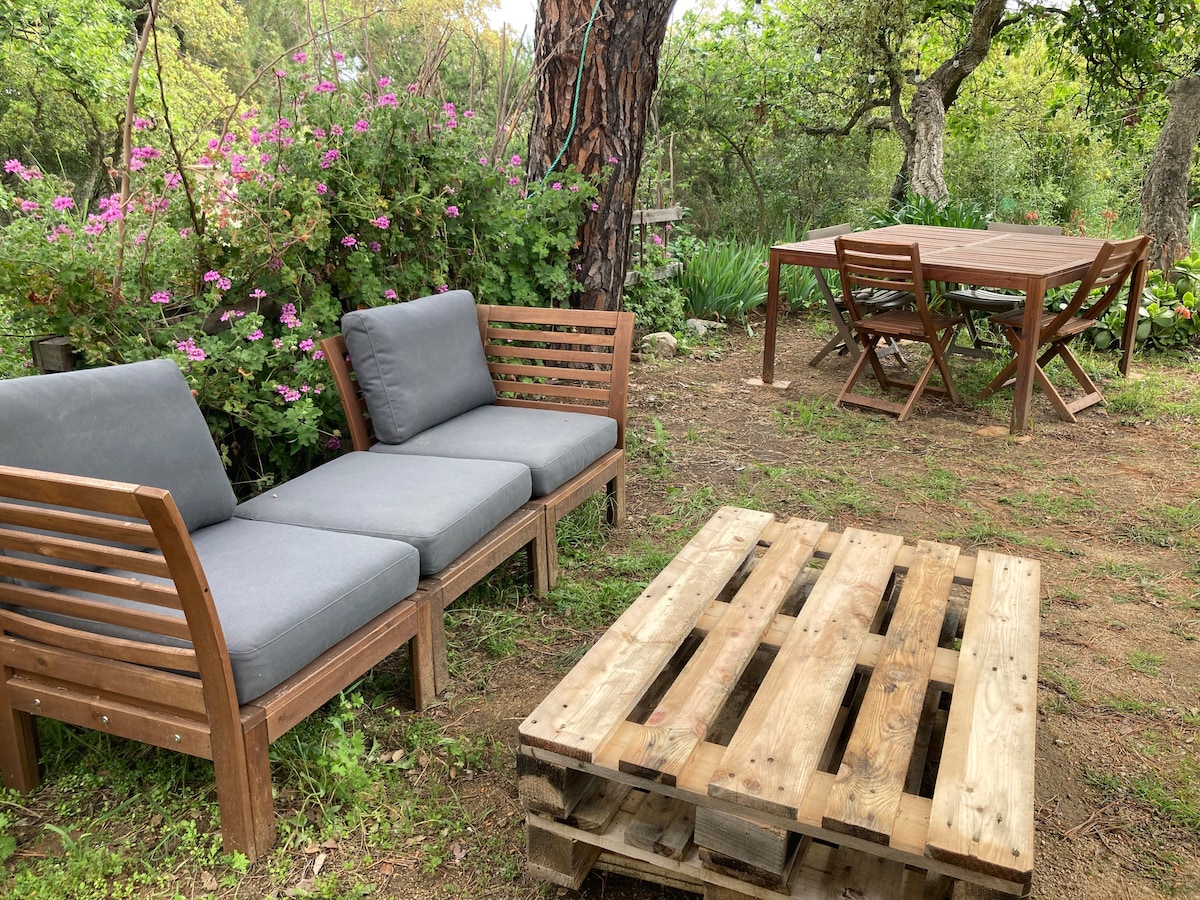 A garden area is presented with wooden furniture, including a sofa with gray cushions and a rustic wooden table made from pallets. Surrounding flowers and greenery create a natural setting, while a dining table is available for outdoor meals.