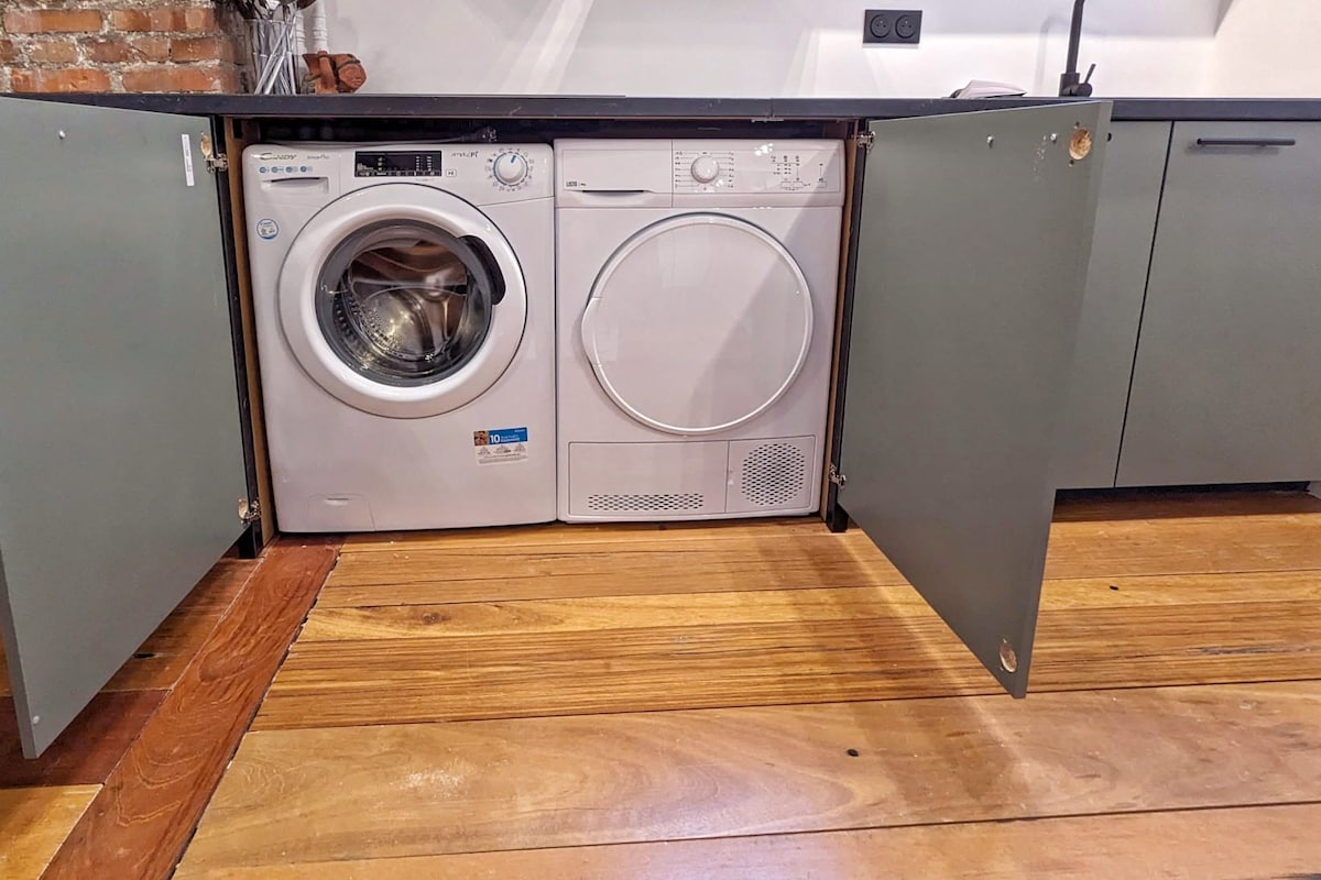 The kitchen features concealed cabinetry, showcasing a washing machine and dryer neatly integrated into the design. The appliances are framed by stylish grey cabinet doors, allowing for a streamlined appearance. The floor displays warm wooden tones, contributing to the rustic charm of the space.