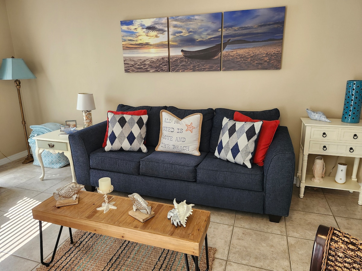 A cozy living area is shown featuring a deep blue sofa adorned with decorative pillows. A wooden coffee table sits in front, complemented by a woven rug and coastal-themed decor. Soft natural light filters through the space, creating a relaxed atmosphere.
