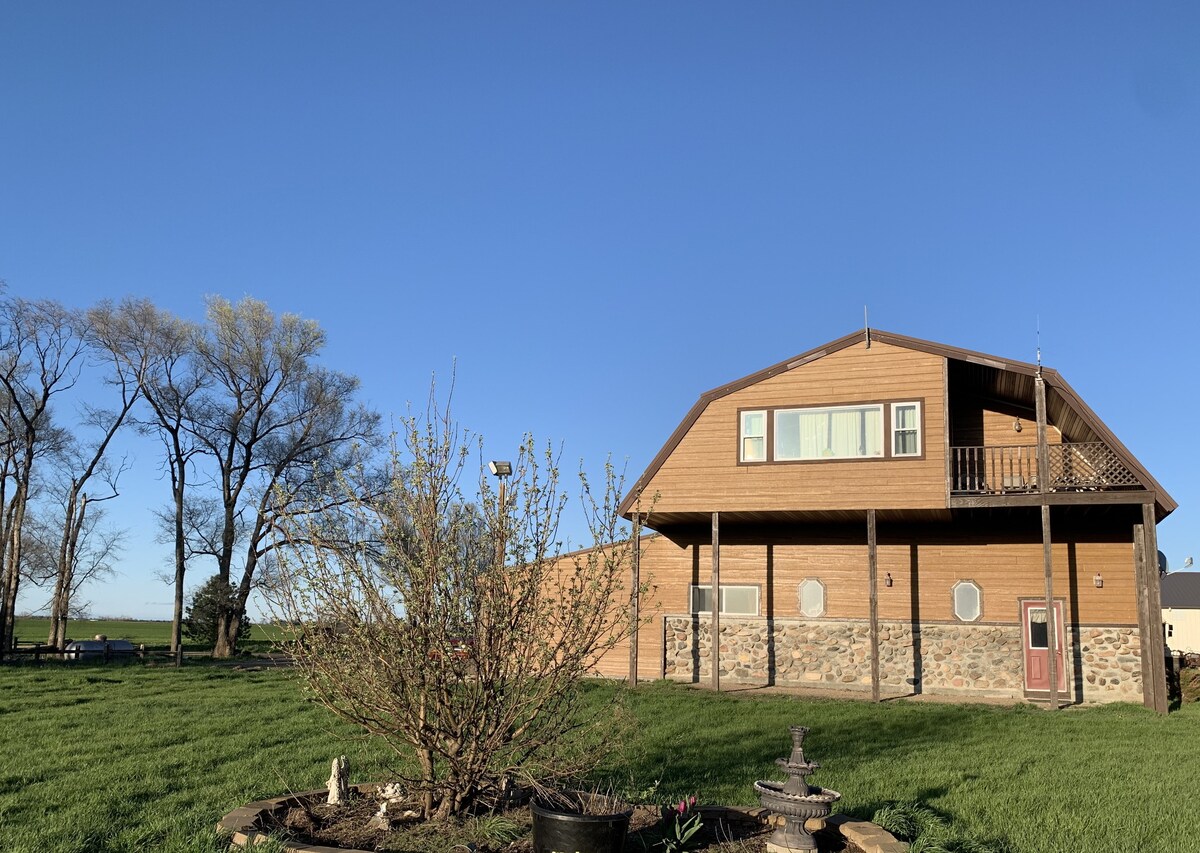 A two-story lodge is seen surrounded by green grass, featuring a combination of wooden and stone exteriors. A balcony extends from the upper level, and several trees are present nearby. The clear blue sky contributes to the serene rural setting.
