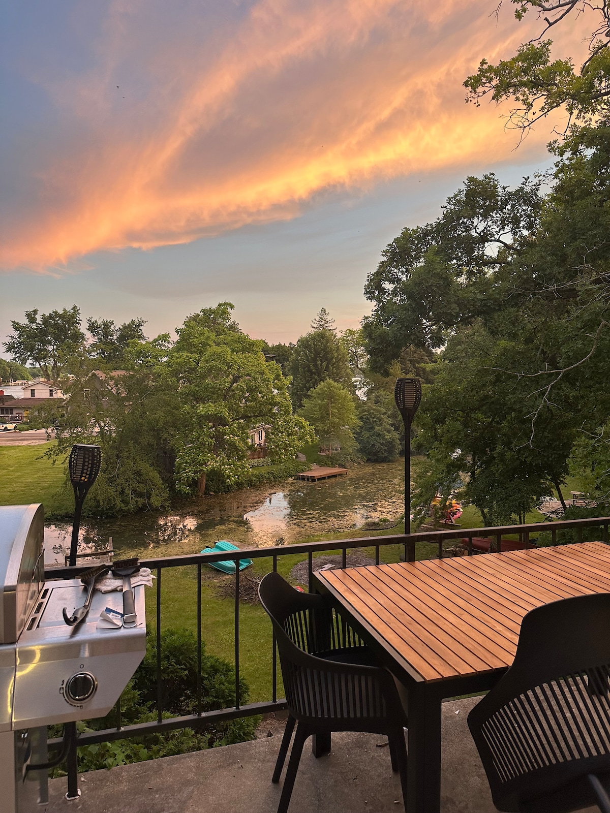 A spacious patio with a wooden dining table and black chairs overlooks a serene yard. A charcoal grill is positioned to one side. In the background, a lush green landscape is visible, along with a tranquil waterway reflecting the soft colors of the sunset.