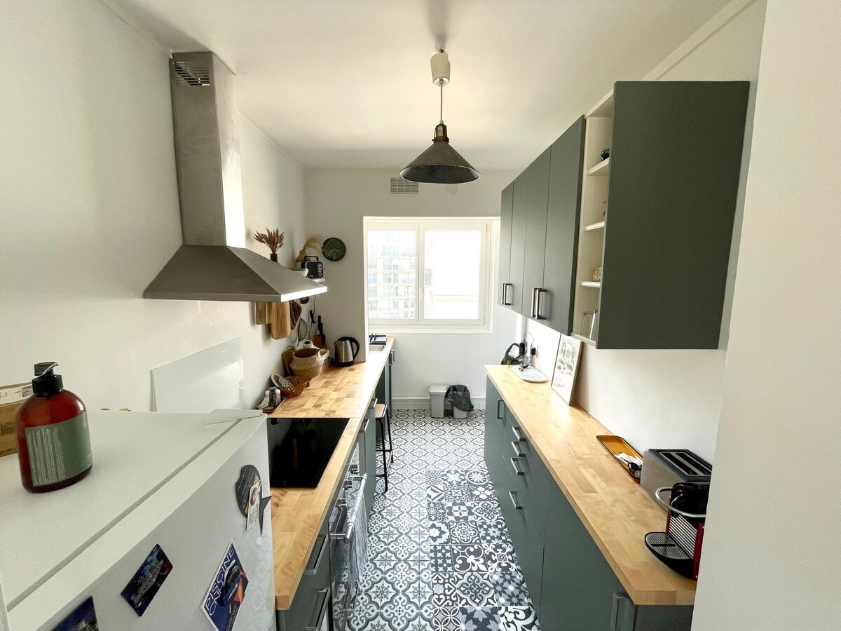 The kitchen showcases a modern design with light-colored cabinetry and wooden countertops. Stainless steel appliances are positioned along one side, while patterned flooring adds character. Ample natural light is provided by the large window, enhancing the space's functionality.