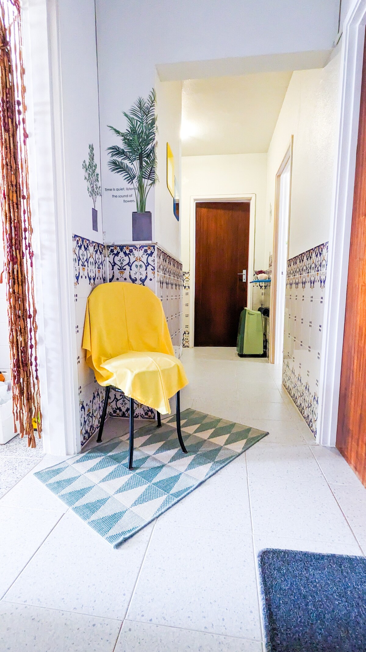 A narrow hallway features a patterned area rug positioned under a minimalist chair draped with a yellow throw. Decorative tiles line the walls, contrasting with the bright, airy space. A door at the end leads to another room, while a potted plant adds a touch of greenery.