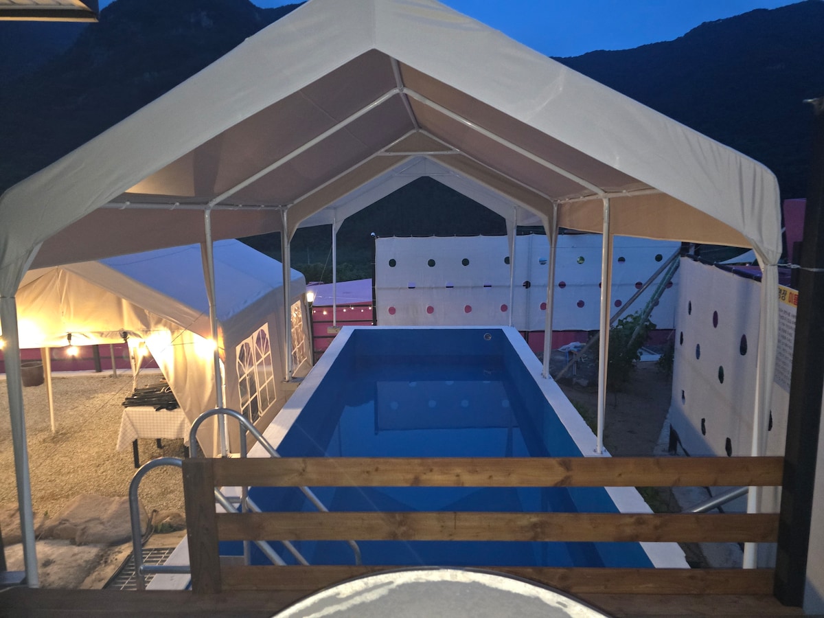An outdoor area features a covered pool and a separate tent structure. The pool, surrounded by a gravel landscape, reflects the evening sky. Colorful dot designs are visible on the nearby walls, enhancing the vibrant atmosphere of the space.