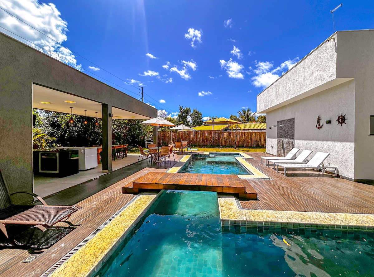 An outdoor leisure area features a spacious pool surrounded by wooden decking. Lounge chairs are positioned for relaxation. A shaded dining space is visible with several tables and chairs. A grill is situated nearby, and bright blue skies frame the scene.