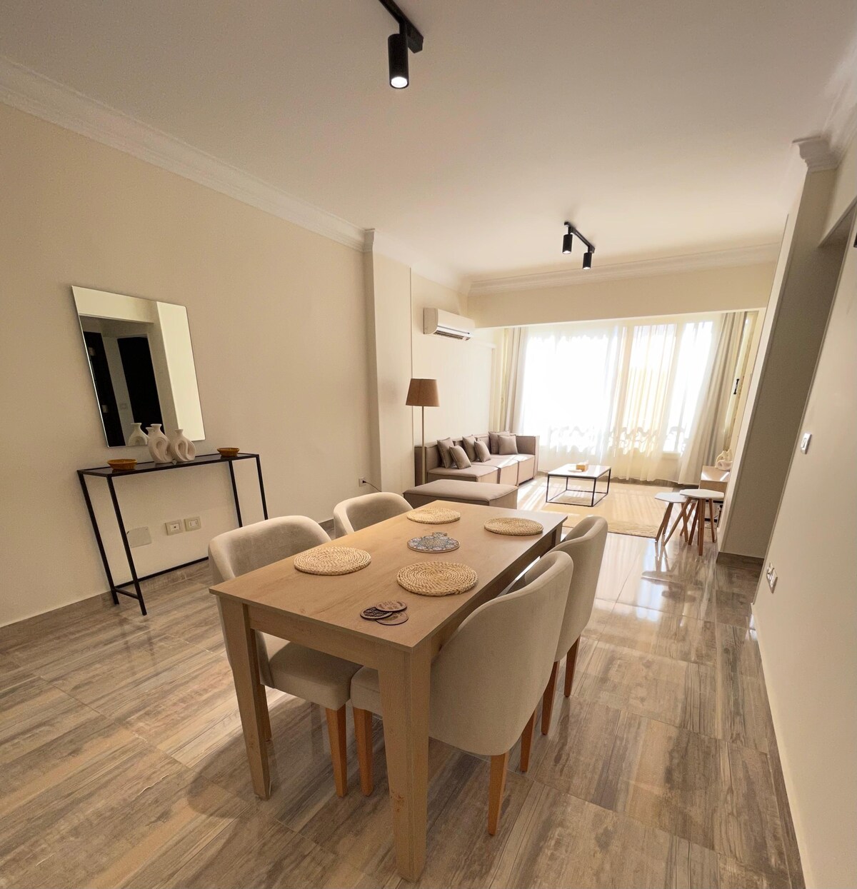 A dining area features a light-colored table surrounded by plush, upholstered chairs. A mirror is positioned on a wall, reflecting natural light that enters from large windows. A comfortable sofa is seen in the background, complemented by soft curtains that frame the view.