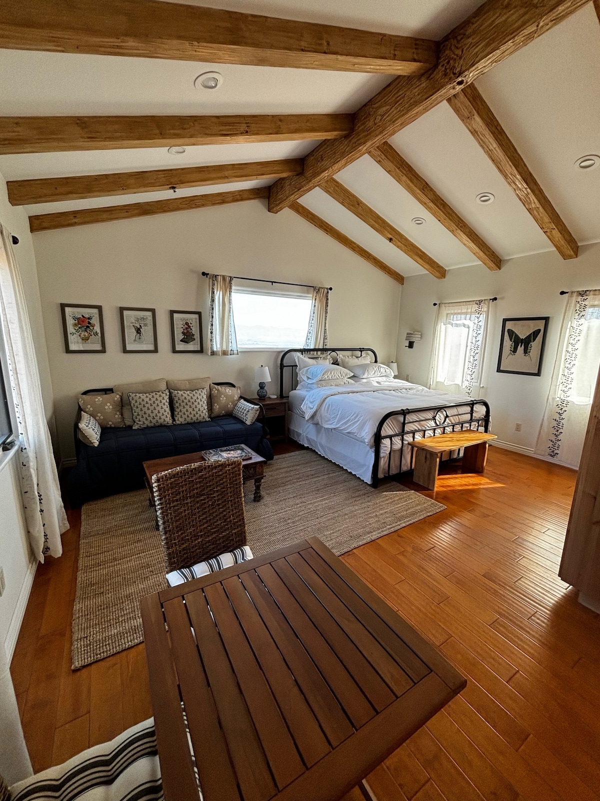 A bright bedroom features a spacious bed with white linens, complemented by decorative pillows. A blue sofa and a small wooden table provide additional seating, while exposed wooden beams add character to the ceiling. Natural light enters through a window dressed with light curtains.