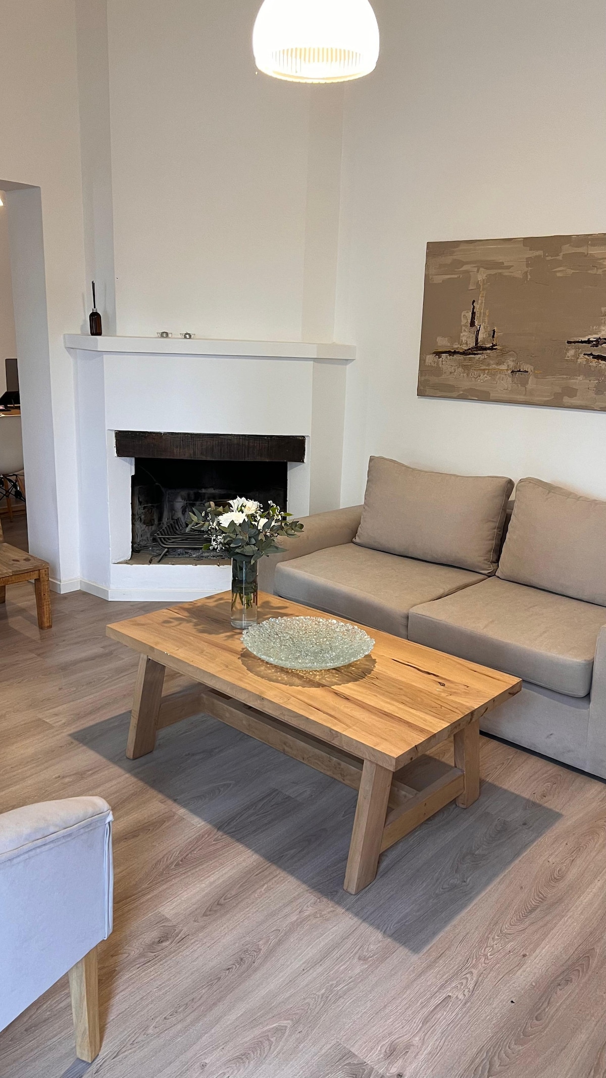 A living area features a light-colored sofa and a wooden coffee table, adorned with a decorative bowl. A fireplace is positioned prominently against the wall, and natural light is supported by a pendant light overhead. A piece of wall art complements the neutral decor.