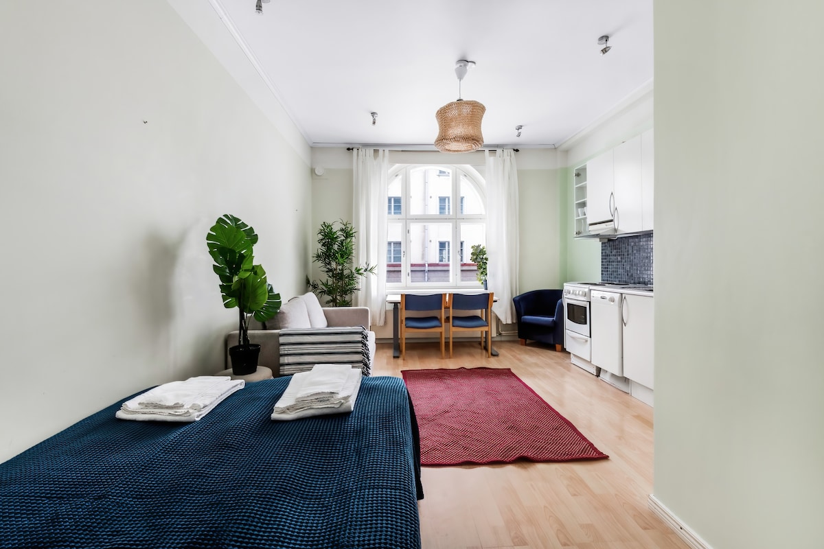 The bright studio features a cozy sleeping area with a blue bedspread and fresh towels. A small dining table with chairs sits adjacent to a light kitchen, which includes ample storage and modern appliances. Two large windows allow natural light to fill the room.