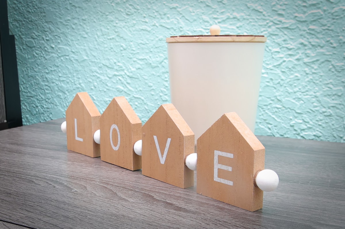 Decorative wooden blocks shaped like houses spell out the word 'LOVE.' They are positioned on a wooden surface alongside a simple, light-colored container. A textured aqua wall serves as a backdrop.