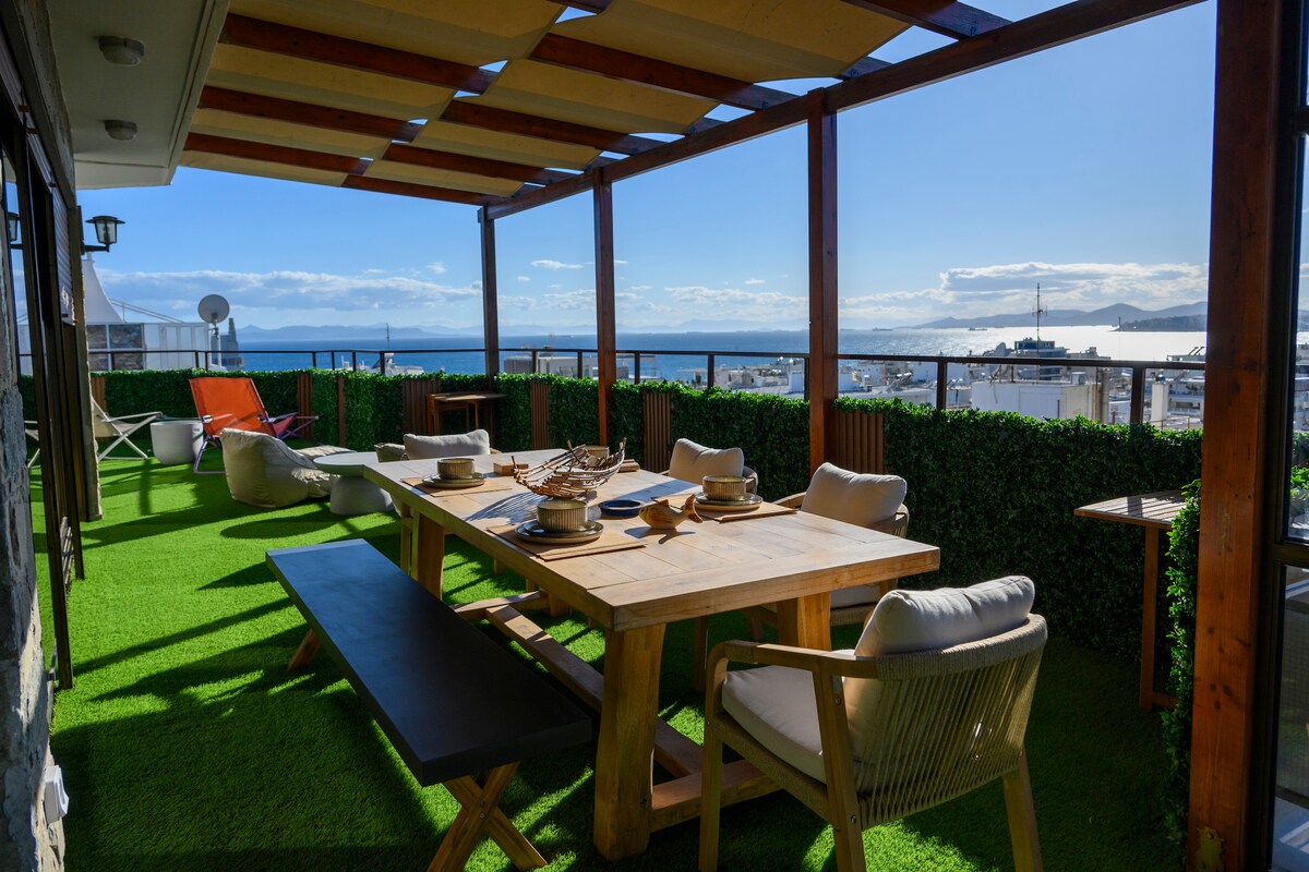 An expansive terrace showcases a wooden dining table, accompanied by cushioned chairs and benches. Lush greenery lines the perimeter, offering privacy. A stunning view of the sea and skyline is framed by the open roof, allowing ample natural light to illuminate the space.