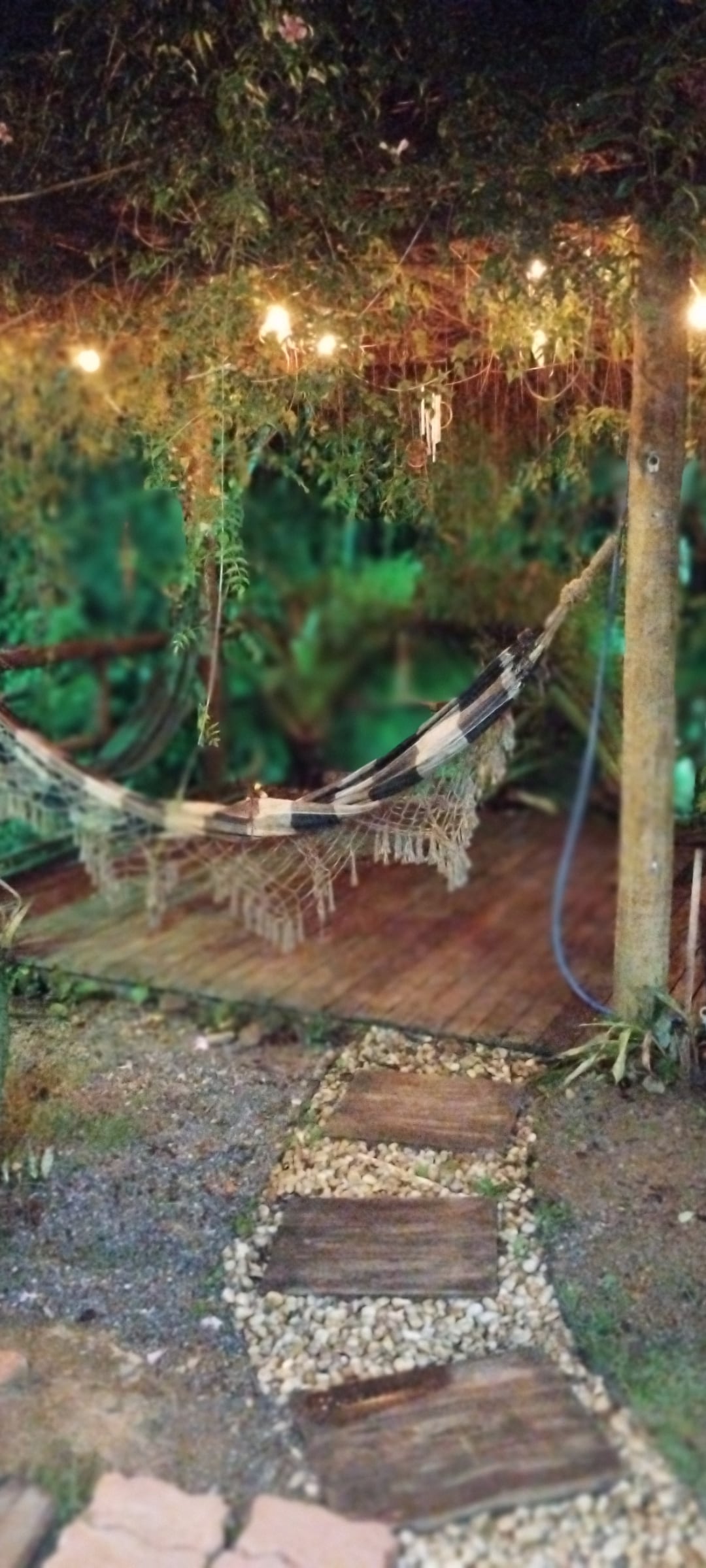 A serene outdoor area is depicted, featuring a comfortable hammock suspended between two trees. Soft lighting is provided by string lights overhead, creating a gentle ambiance. A pathway made of wooden planks and pebbles leads toward the seating space, surrounded by lush greenery.