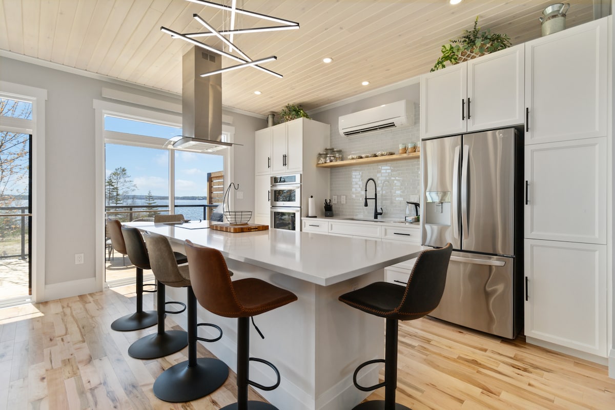 A modern kitchen features a large island with four high-backed stools. Sleek white cabinetry contrasts with stainless steel appliances, including an oven and refrigerator. Natural light floods the space through a sliding glass door, highlighting the minimalist design and elegant finishes.