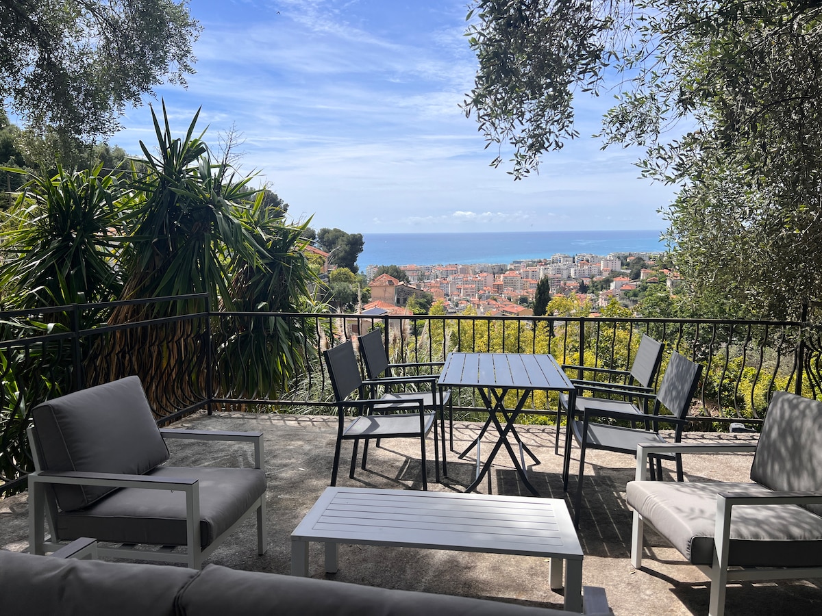 An outdoor terrace is visible, showcasing several seating arrangements, including a table with four chairs and lounge chairs. Lush greenery frames the space, offering views of the sea and surrounding landscape. The sky is clear, enhancing the serene atmosphere.