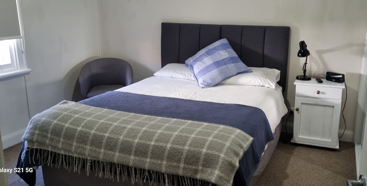 A modern bedroom features a spacious bed with a neatly arranged blue and white checked pillow. A soft blanket drapes over the bed, and a small bedside table holds a lamp and clock. A comfortable chair is positioned nearby, with natural light coming through a window.