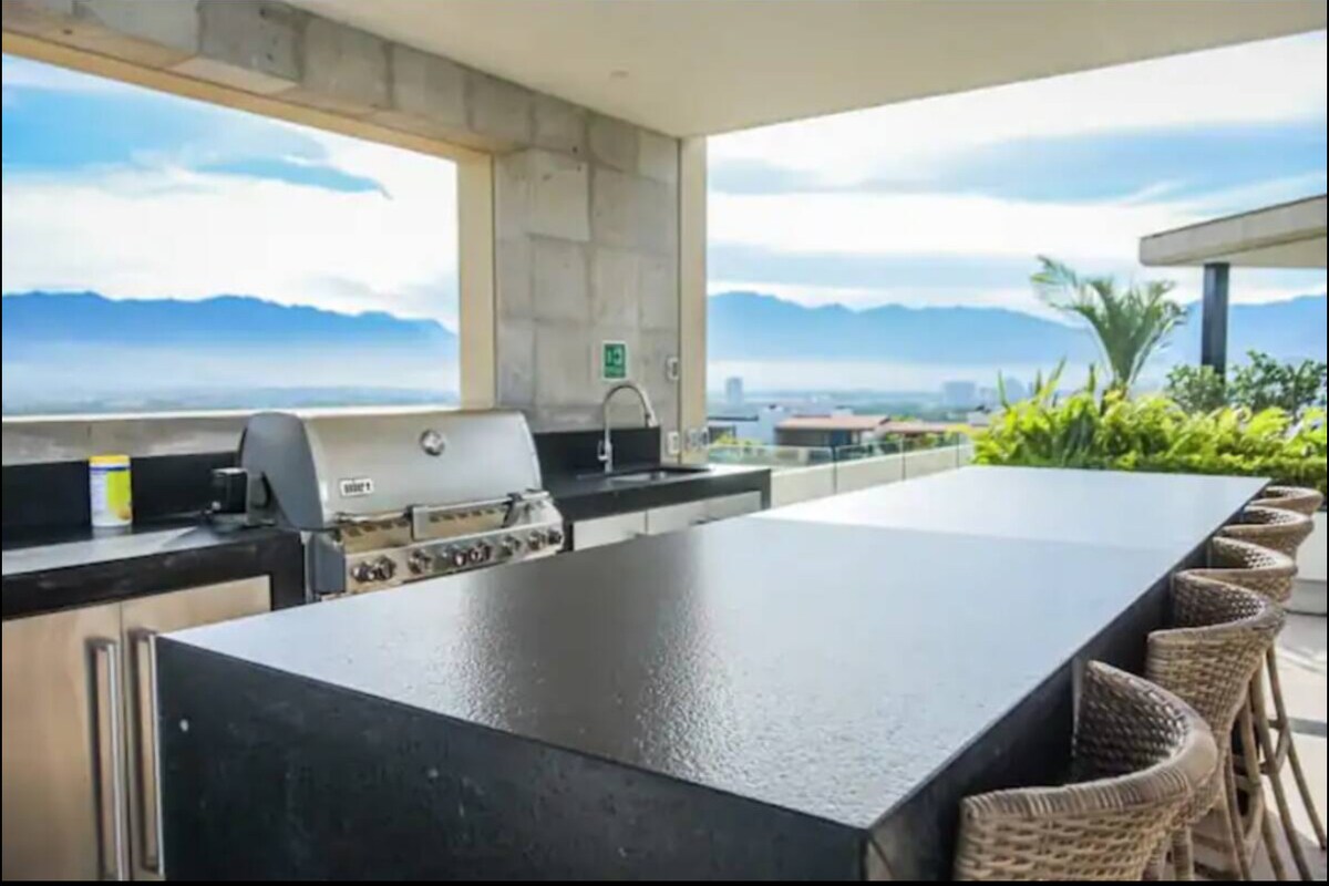 An outdoor kitchen area features a large black countertop with seating for five, alongside a modern barbecue grill. A panoramic view of the mountains and surrounding landscape is visible through a large window, enhancing the open-air design.