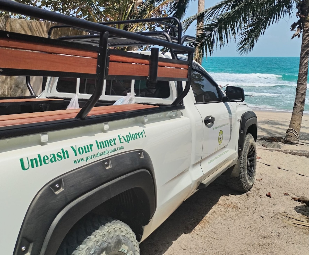 A rugged 4x4 vehicle is parked on sandy beach terrain, with palm trees surrounding it. The ocean can be seen in the background, enhancing the sense of adventure. The side of the vehicle features a logo and a slogan encouraging exploration.
