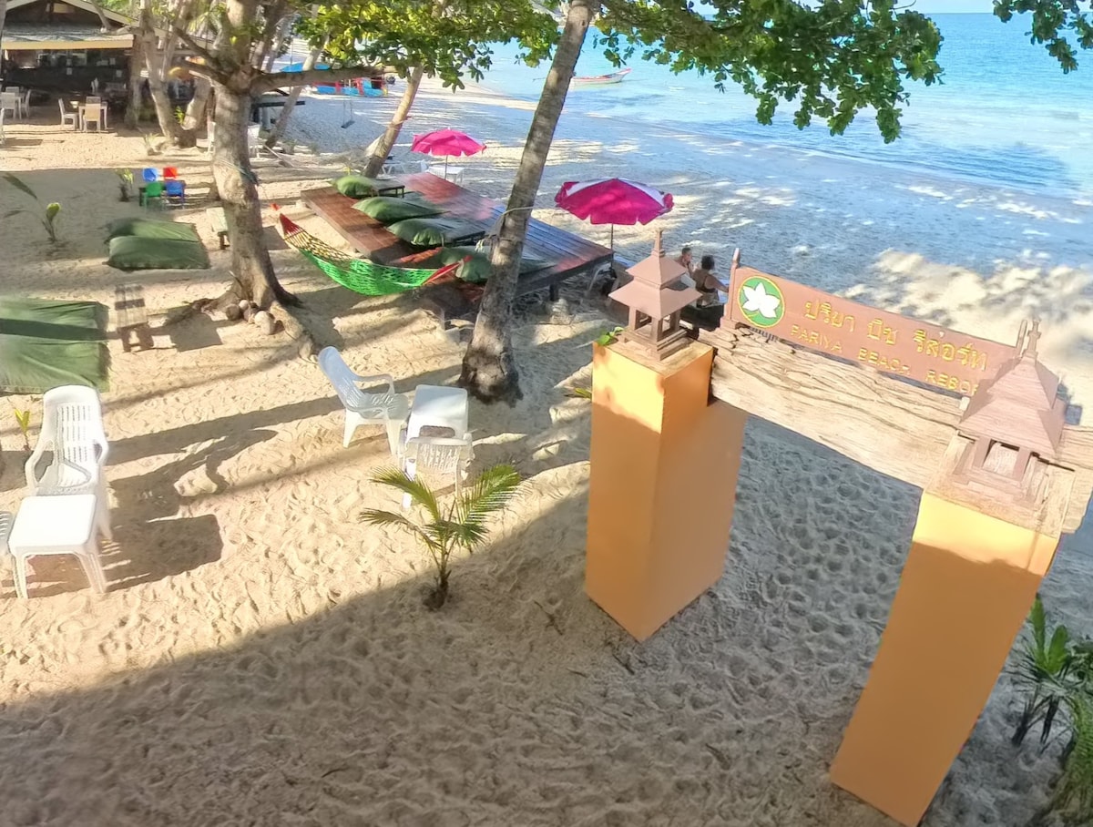 The beach area features white sand framed by greenery, with several colorful hammocks under tall trees. A welcoming sign stands at the entrance, marked with a logo and text in Thai. White plastic chairs and tables are scattered nearby, inviting relaxation by the shore.