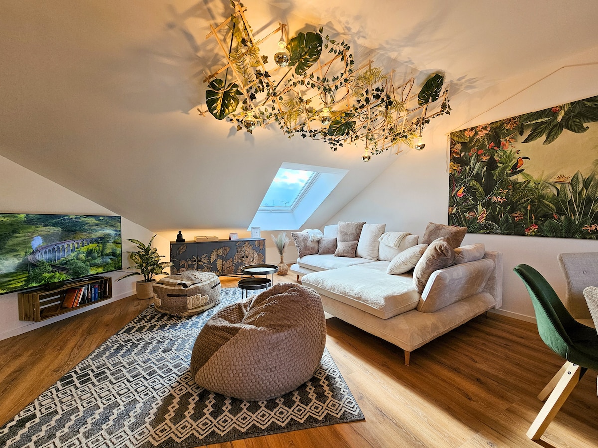 The living area features a spacious sectional sofa adorned with various cushions, inviting comfort. A large television sits on a wall-mounted shelf beside a decorative plant. Natural light streams through a skylight, illuminating a colorful wall mural and a textured rug that adds warmth to the wooden floor.