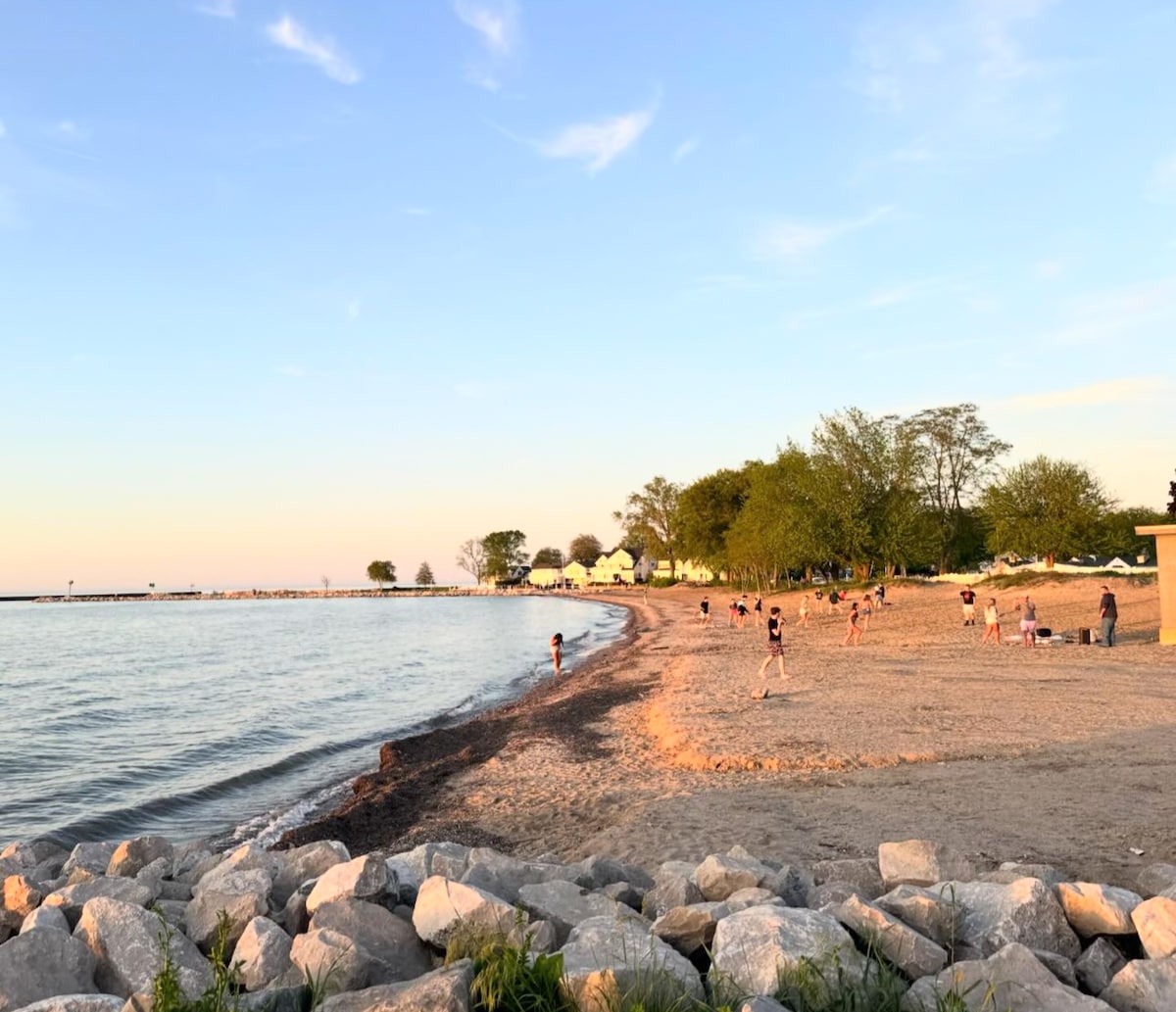 A sandy beach extends along the shoreline, bordered by smooth rocks. Gentle waves lap at the shore, with several individuals walking and enjoying the pleasant weather. Green trees provide shade in the background, creating a calming natural environment.