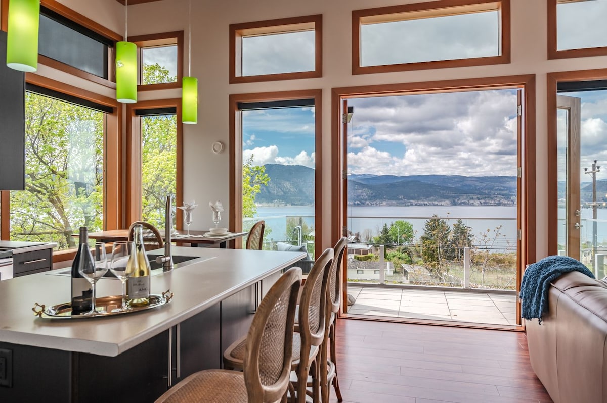 A spacious kitchen features modern fixtures and light wood cabinetry. Large windows provide expansive views of Okanagan Lake and surrounding landscapes. Natural light fills the area, while green pendant lights add a touch of color. A dining space is visible, suitable for enjoyable meals.