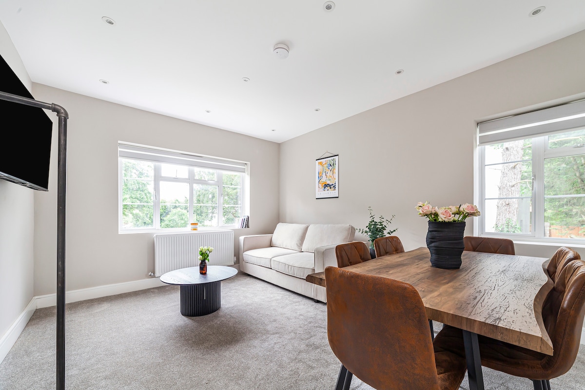 A spacious living area is illuminated by natural light from large windows. A neutral-colored sofa is positioned across from a flat-screen TV. A circular coffee table rests in the center, surrounded by a dining table with four leather chairs. A potted plant and flowers add a touch of decor.