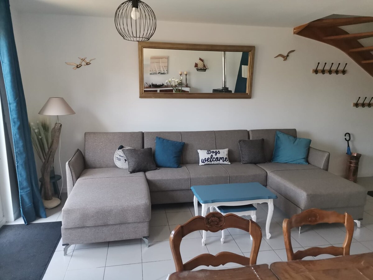 A spacious living area is presented, featuring a modern gray sectional sofa with accent pillows. A light blue coffee table complements the seating. Natural light streams through large windows, enhancing the room’s openness. A dining table is visible in the background.