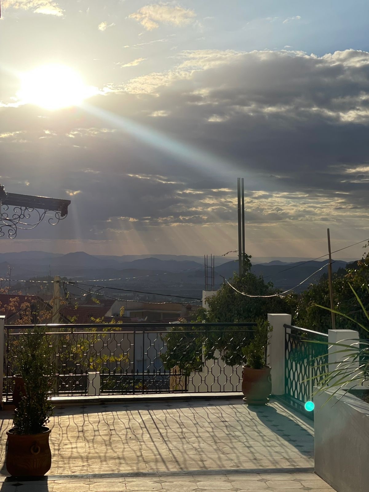 A sunlit terrace is shown, featuring a railing that overlooks rolling hills and valleys. The sky hosts a mix of clouds with rays of sunlight streaming through. Potted plants are placed along the edge, complementing the tranquil outdoor space.