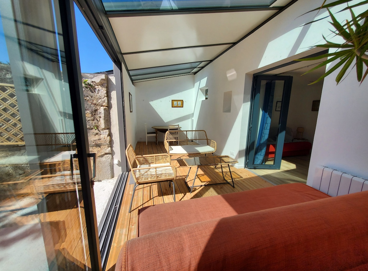 The image shows a bright and airy veranda featuring a small seating area consisting of two armchairs and a table. Large glass doors provide a view into the adjoining space, where a red bed is partially visible. Sunlight fills the area, highlighting the wooden floor.