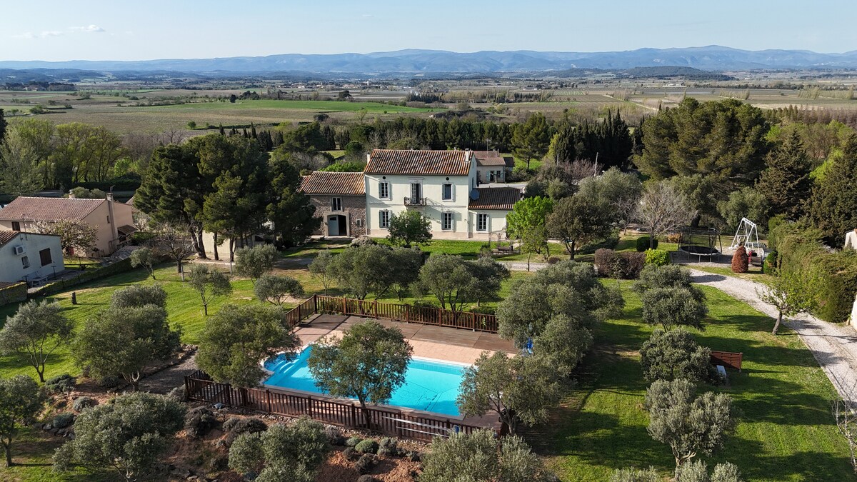 Aerial view of the property showcases the extensive green grounds surrounding the large heated pool. Olive trees are scattered throughout the landscape, with rolling hills visible in the background under a clear sky.