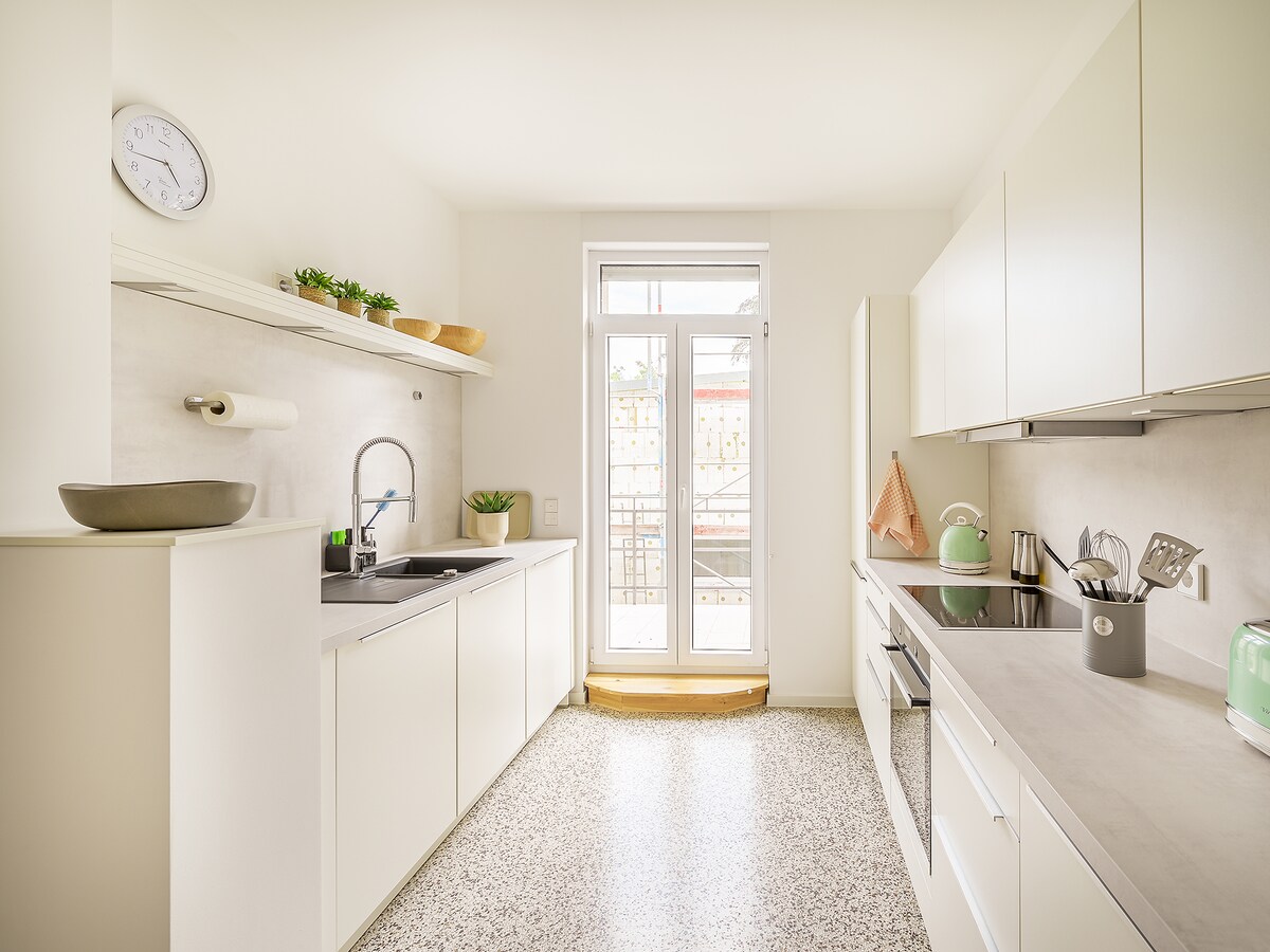 The kitchen is bright and modern, featuring sleek white cabinets and a spacious counter. Natural light floods in from the glass doors leading to a small balcony. Appliances such as a kettle and oven are visible, alongside potted plants that add a touch of greenery.