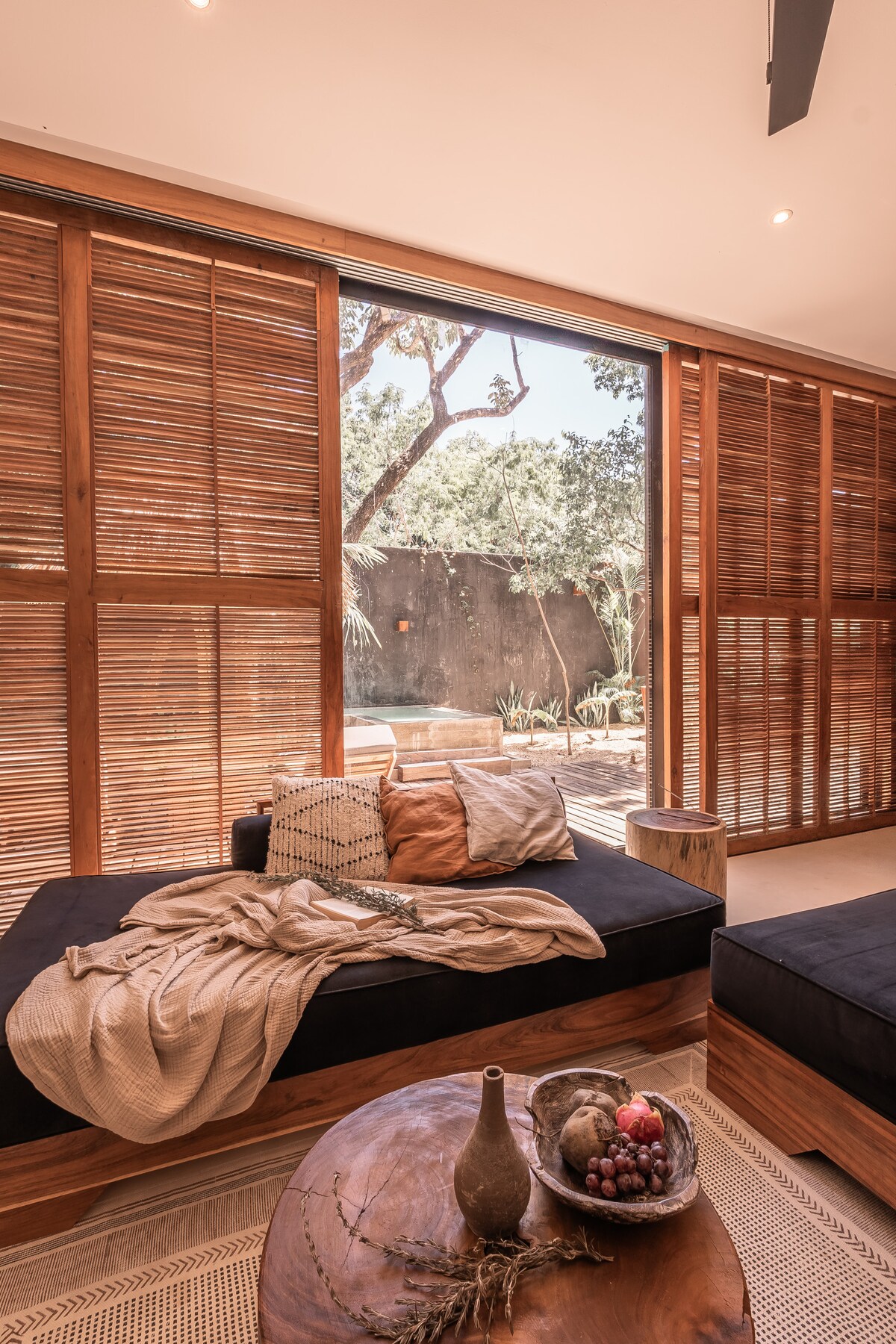 A spacious seating area features dark upholstered sofas layered with a neutral throw and decorative pillows. Natural light filters through wooden slatted shutters, revealing a glimpse of greenery outside. A round wooden coffee table displays a bowl of fruit, adding a touch of warmth.