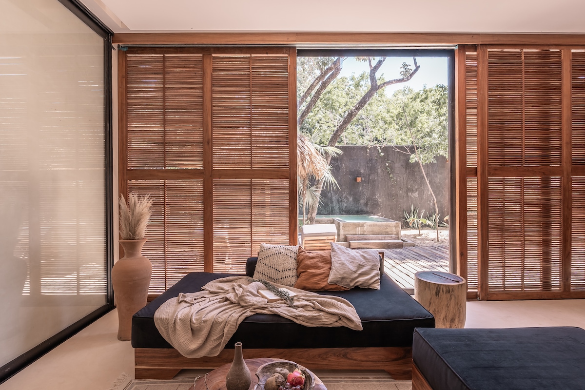 A comfortable seating area is present, featuring dark cushions and a light throw blanket over a stylish couch. Wooden sliding doors reveal a natural outdoor space with trees and a sunlit patio area, enhancing the connection to the surrounding greenery.