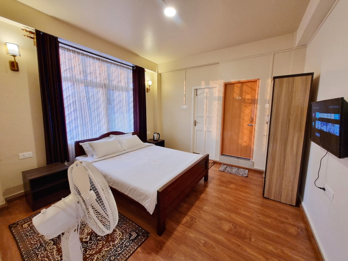 A well-lit bedroom features a spacious bed with a minimalist design. A standing fan is positioned nearby, and a wooden cabinet adds storage space. Natural light enters through large windows with sheer curtains, creating a calming environment. A TV is mounted on the wall opposite the bed.