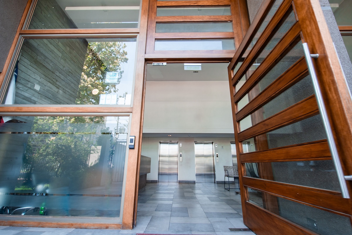 A modern entrance is displayed, featuring large glass doors framed in warm wood. The spacious lobby showcases stone flooring, with two elevators visible in the background. Natural light filters through, highlighting the greenery outside.