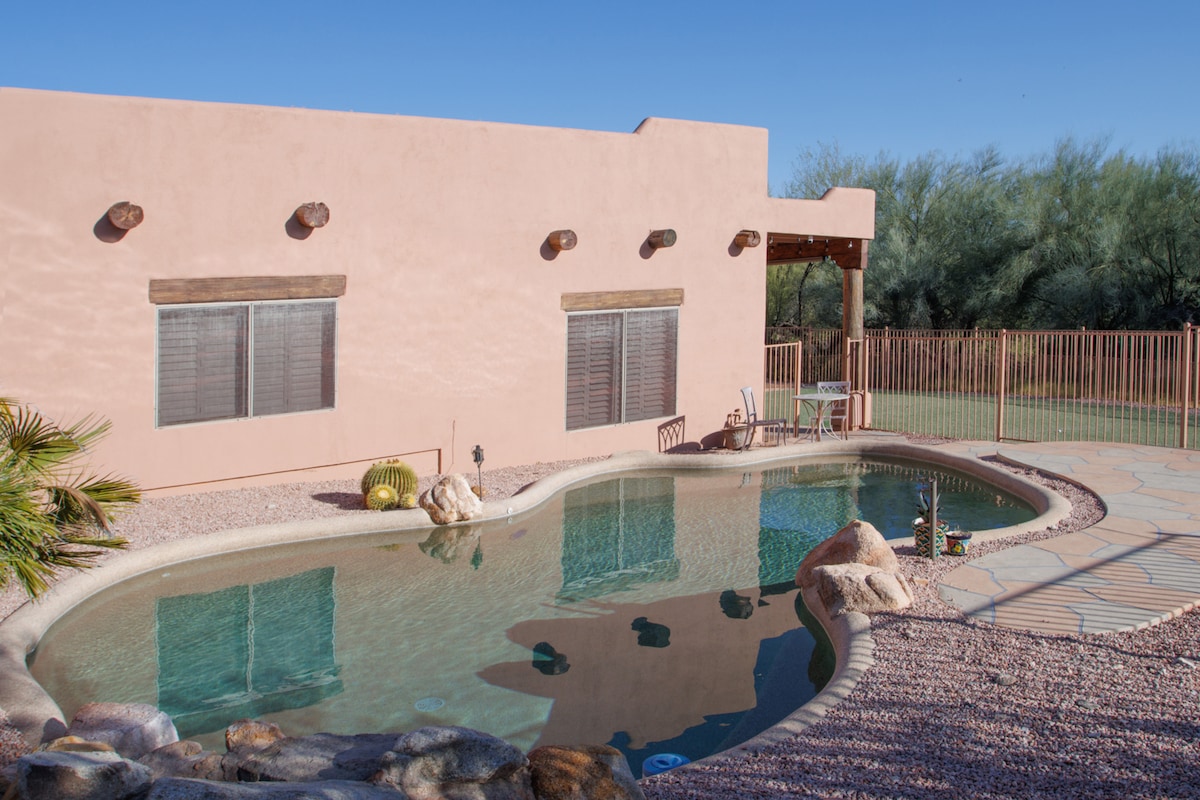 A heated pool is featured in a private outdoor space, surrounded by smooth stones and arid landscaping. The pink adobe-style exterior of the home reflects in the clear water. A shaded seating area is visible beside the pool, enhancing outdoor relaxation.