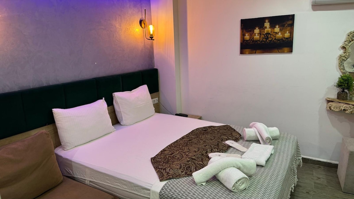 A cozy sleeping area features a spacious bed with a decorative throw blanket. Plush pillows are arranged neatly at the headboard. Soft lighting is provided by wall sconces, and a piece of artwork depicting a cityscape is displayed on the wall.