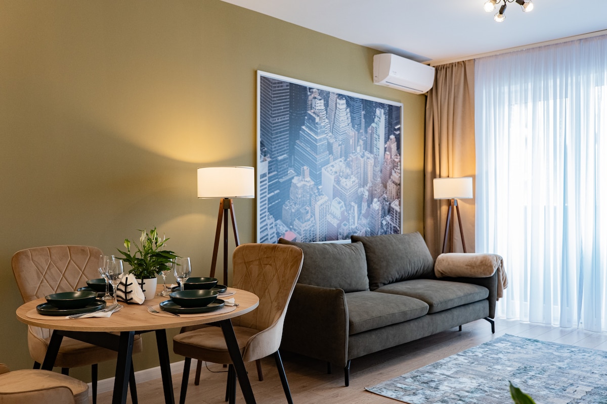 A living area is presented with a soft brown sofa and a round dining table set for four. A large cityscape artwork adorns the wall. Soft lighting from two floor lamps and natural light filters through sheer white curtains, creating a comfortable atmosphere.