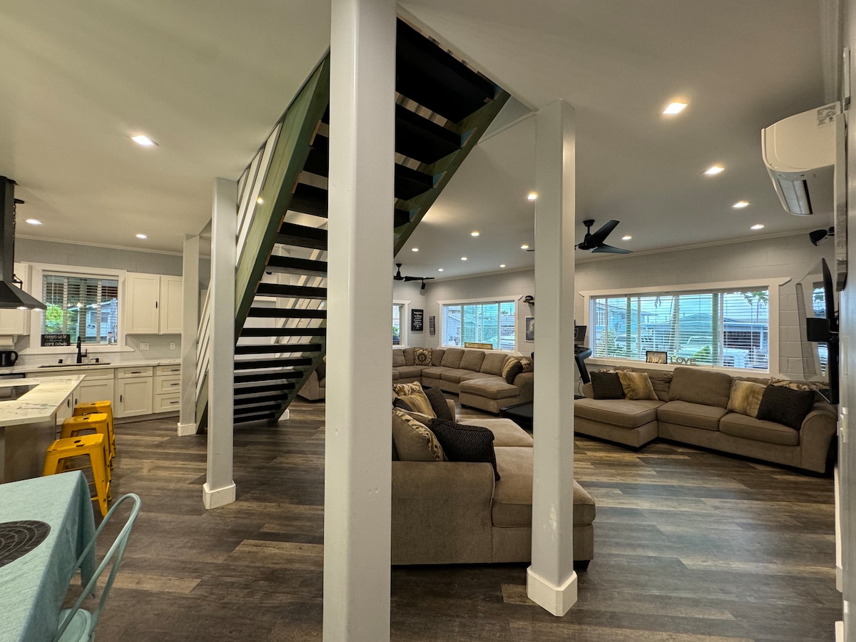 An open living space is highlighted by a sectional sofa arranged around a coffee table. Natural light floods the area through multiple windows. A modern kitchen with white cabinetry is visible, complemented by a staircase leading to the upper floor. Bright yellow stools add a splash of color.