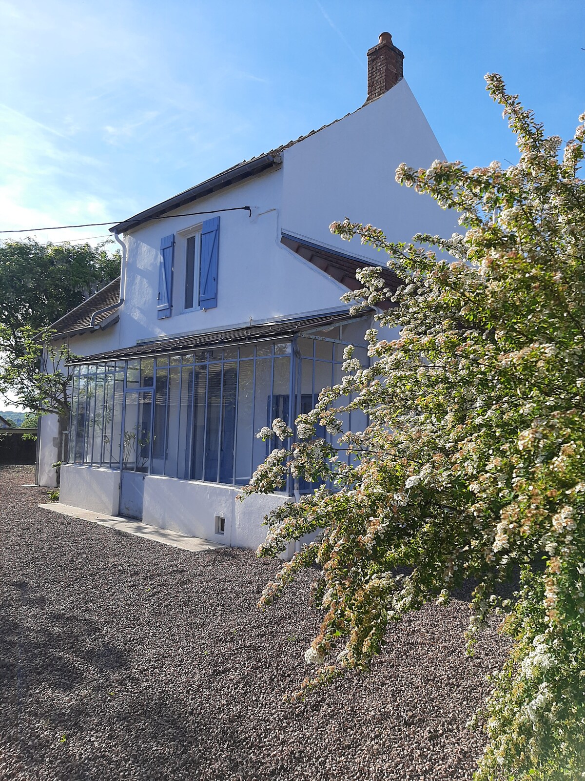 The exterior of a freshly renovated house is shown, featuring traditional blue shutters and a charming glass-enclosed porch. A flowering shrub adds a touch of greenery to the gravel pathway leading to the entrance, while the clear sky enhances the house's inviting façade.
