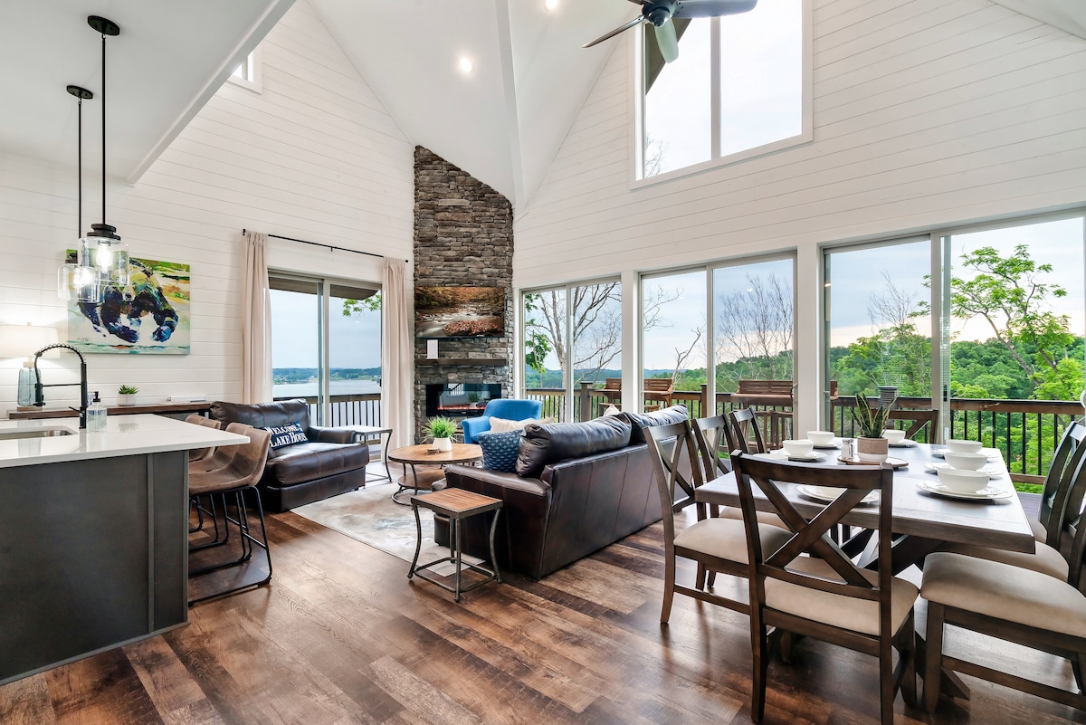 An open living area is highlighted by tall windows offering views of the surrounding nature. A stone fireplace anchors the space, complemented by comfortable seating. A dining table for eight sits nearby, with an adjacent kitchen bar that allows for casual dining.